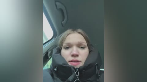 Media: A video of a young woman with fair skin and light brown hair, wearing a black jacket, sitting in a car. The car's interior is dimly lit, with a window partially visible.