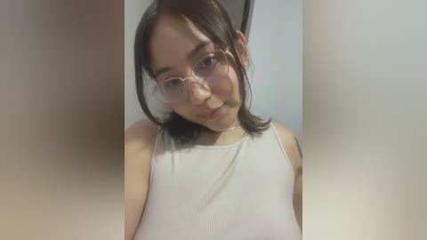 Media: Video of an Asian woman with straight dark hair and glasses, wearing a white ribbed tank top, in a minimalistic setting with blurred background.