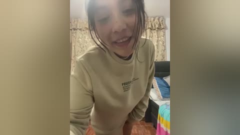 Media: A video of a young Asian woman with wet hair, wearing a beige sweatshirt, smiling while leaning over a brown blanket in a cluttered bedroom with patterned curtains and a bed.