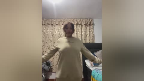 Media: Video of a woman with medium skin tone and brown hair, wearing a beige sweater, standing in a cluttered bedroom with floral curtains, bed, and clothes on the floor.