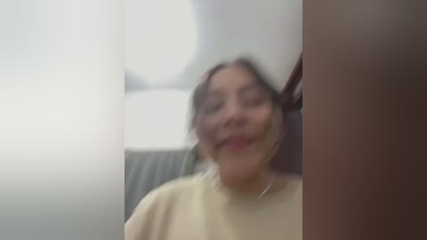 Media: A blurred video of a woman with medium skin tone, long dark hair, and a light yellow top, smiling in a room with a white wall and a lamp.