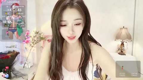 Media: A video of an Asian woman with long, straight black hair, wearing a white tank top, sitting in a brightly lit room with a floral arrangement and a table lamp in the background.