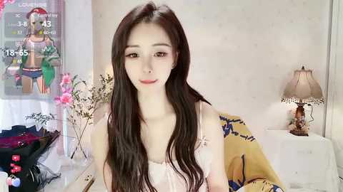 Media: Video of a slim, young Asian woman with long, straight black hair, wearing a white tank top, smiling in a cozy bedroom with a lamp, pink flowers, and a bed with a yellow sheet.