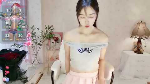 Media: Video of a slender East Asian woman with long dark hair, wearing a light gray off-the-shoulder crop top with \"TINARAH\" and pastel pink pleated skirt, sitting in a white chair. Background includes a digital TV displaying a game, a table lamp, and a floral arrangement.