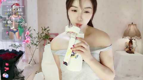 Media: A video shows an Asian woman with straight brown hair, drinking from a milk bottle, wearing a light grey off-shoulder top. The room has pastel walls, a lamp, and a TV showing a live stream.