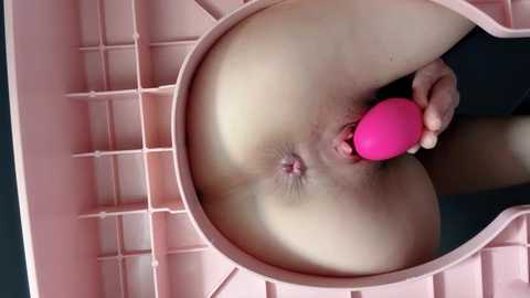 Video of a nude person with fair skin, holding a pink vibrator against their exposed genitalia. The background features a pink, grid-like plastic box.