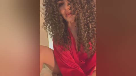 Media: Video of a woman with light brown skin and voluminous, curly hair, wearing a red blouse, seated indoors with a warm, neutral background.