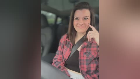 Video of a smiling woman with shoulder-length brown hair, wearing a red plaid shirt and black tank top, driving a car with a seatbelt on.