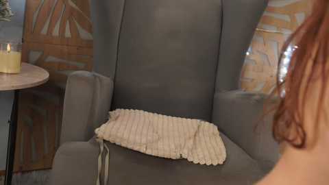 Media: Video of a cozy, gray armchair with a cream knitted pillow, set against a rustic wooden backdrop and a lit candle on a small round table.