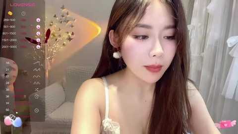 Media: A video of an Asian woman with long, straight brown hair, wearing a white lace bra, standing in a well-lit, modern bedroom with a bed and white curtains.