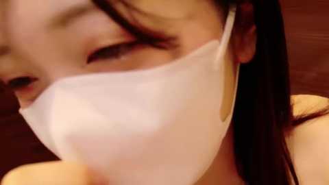 Video of an Asian woman with long dark hair, wearing a white surgical mask, her eyes closed, focusing on her face, blurred background.