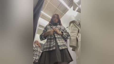 Video of a young woman in a fitting room, wearing a plaid shirt over a black skirt, adjusting her outfit. The room has white walls, beige carpet, and hanging clothes.