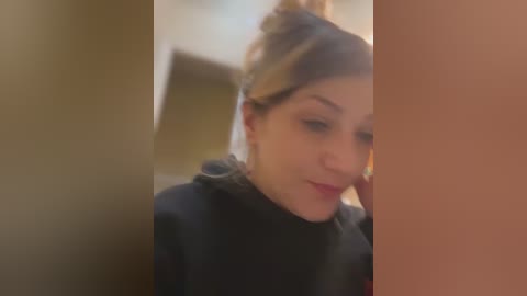 Media: A video of a young woman with light skin and brown hair tied in a bun, wearing a black turtleneck sweater, slightly blurred and out of focus.