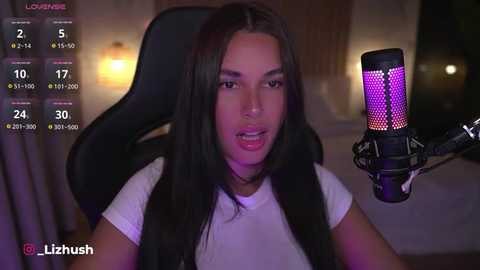 Media: Video of a young woman with long, dark hair and a light complexion, wearing a white top, sitting in a black gaming chair. She's using a purple-lit microphone, with a live stream overlay showing her stats.