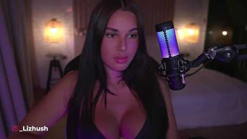 Video of a dark-haired woman with long, straight hair, wearing a black top, singing into a microphone in a dimly lit room with soft lighting and a bed visible in the background.