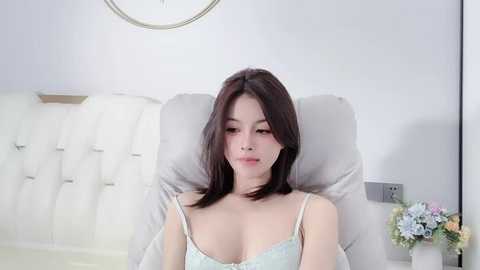 Video of an Asian woman with straight, dark hair, wearing a light blue, lace-trimmed bra, reclining on a white, tufted headboard in a minimalist, light-colored room.