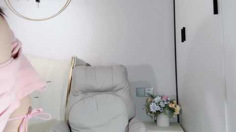 Media: A video of a minimalist bedroom with a beige recliner, light-colored walls, a round clock, and a small table holding a bouquet of flowers.