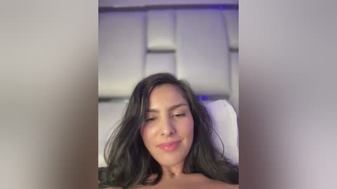 Media: Video of a smiling Latina woman with long, wavy black hair, light skin, and a small nose ring, lying on a white bed with a modern, padded headboard.