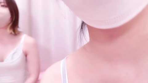 Media: Video of a woman in a white mask and white lace top, seen from the side, with a blurry reflection in the background. Soft, pastel-colored light creates a serene atmosphere.