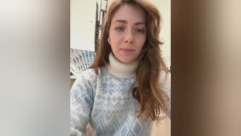 Media: Video of a young woman with long, wavy brown hair wearing a light blue, patterned sweater, standing in front of a beige wall with a wooden chair in the background.