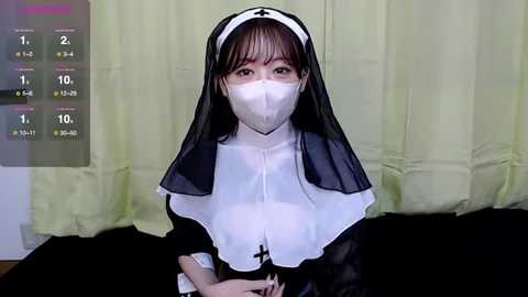 Media: A video of an East Asian woman dressed as a nun, wearing a white mask, black veil, and habit, against a green curtain background.