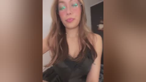 Media: Video of a light-skinned, young woman with long, wavy brown hair, wearing a black satin camisole. Her makeup includes green eyeshadow and pink lipstick. The background is blurred, with neutral tones.