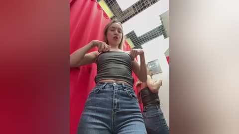 A low-angle video of a young woman with light skin, wearing a grey crop top and high-waisted jeans, adjusting her bra straps. She stands in a brightly lit fitting room with red curtains and white ceiling tiles.