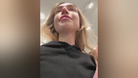 Media: A close-up video of a blonde woman with fair skin, wearing a black dress, taken from a low angle, emphasizing her facial features and neck.