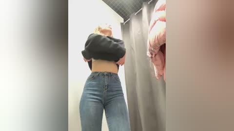 Media: Video of a blonde woman in a changing room, lifting her black sweater to reveal a white midriff, wearing high-waisted blue jeans. Background shows beige curtain and pink jacket on a hanger.