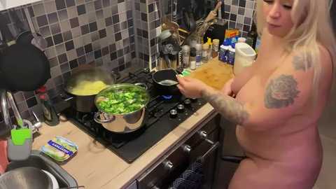 Media: Video of a nude, blonde, tattooed woman cooking in a small kitchen with a tiled backsplash, stainless steel pots, and utensils.