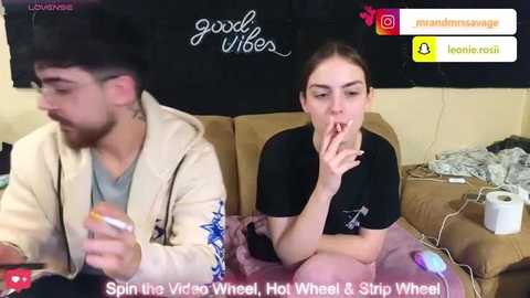 Video of a young woman with light skin and brown hair, wearing a black shirt, sitting on a beige couch, blowing a vape pen. Background shows a black wall with \"good vibes\" text.