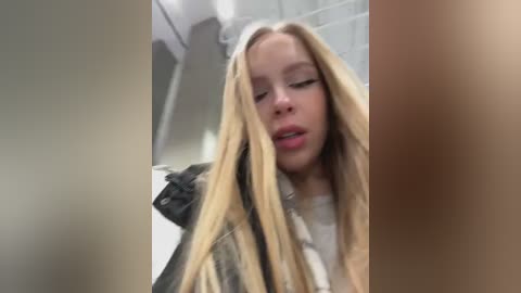 Media: A blurry video of a young woman with long blonde hair, wearing a black leather jacket and a white shirt, looking slightly disheveled in a bathroom with a tiled wall and shower.