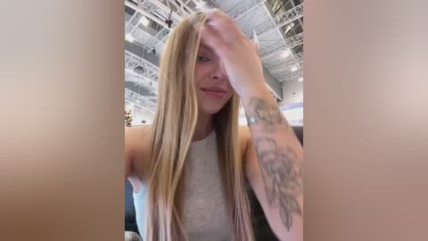 Video of a young woman with long, straight, blonde hair, wearing a sleeveless gray top, sitting in a modern, brightly-lit room with visible ceiling beams. She has a tattoo on her left arm and is touching her hair.