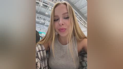 Video of a young Caucasian woman with long blonde hair, wearing a grey tank top and plaid shirt, pouting in a brightly lit indoor venue with high ceilings and exposed beams.