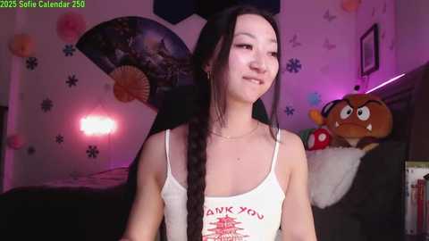 Media: A young Asian woman with long black hair and a slender build, wearing a white tank top, smiles in a cozy room with decorative elements and a plush toy.