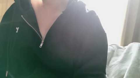 Media: Video of a person's upper torso in a black zip-up hoodie, standing indoors with a blurred background featuring beige curtains and a bed with light-colored bedding.