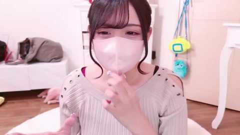 Media: Video of a young Asian woman with dark hair, wearing a white mask, sitting on the floor in a brightly lit room, holding a pink dildo.