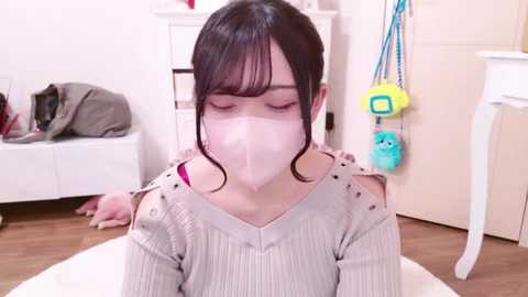 Media: Video of an Asian woman with short black hair, wearing a beige sweater, white mask, and blue gloves, kneeling on a white mat in a clean, white-walled room with wooden floors and toys hanging on the wall.