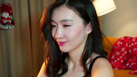 Media: Video of an Asian woman with long black hair, wearing a black tank top, smiling, in a cozy, dimly lit room with a red floral blanket, beige curtains, and a lamp.