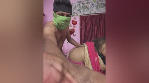 Media: A video shows a shirtless man with a green mask and a woman in pink lingerie and a gold headband, in a pink room with floral wallpaper.