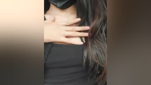 Video of a person with long, dark hair, wearing a black mask and black top, hands on chest, blurred background.