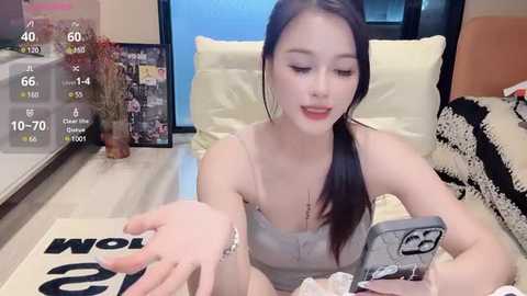 Media: Video of an East Asian woman with long black hair, fair skin, and red lipstick, wearing a light grey spaghetti-strap top, sitting on a beige couch, taking a selfie with a smartphone, with a background featuring a framed photo and a plant.