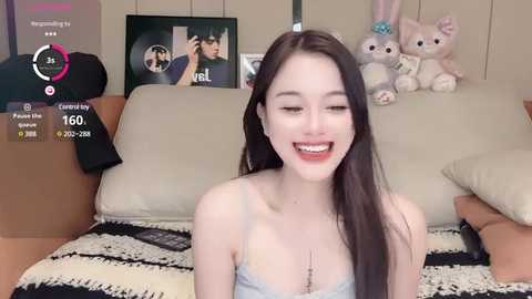 Media: Video of a smiling Asian woman with long black hair, light skin, and a slim figure, wearing a light blue camisole, sitting on a beige couch in a cozy room with stuffed animals and a TV screen displaying a video call.