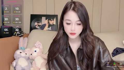 Media: Video of a young Asian woman with long, straight brown hair, fair skin, and a slender figure, wearing a black leather jacket and seated on a beige couch. Background includes a TV displaying a weather forecast and a plush bunny toy.