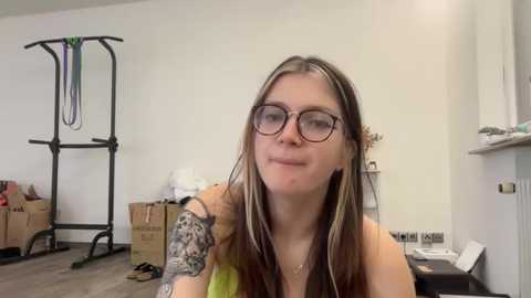 Media: Video of a young woman with long brown hair, wearing glasses, a black top, and a tattooed arm, standing in a cluttered, sparsely furnished room with a metal drying rack, cardboard boxes, and a white wall.