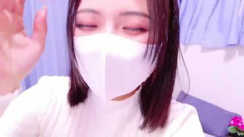 Media: Video of an East Asian woman with straight black hair, wearing a white surgical mask, raising her hand, in a medical setting with blue curtains.