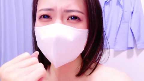 Media: Video of an Asian woman with shoulder-length black hair, wearing a white medical mask, holding a pen in a hospital setting with blue curtains and a blue shirt in the background.