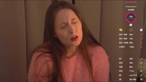 A video of a young Caucasian woman with long brown hair, wearing a pink sweater, eyes closed, and mouth slightly open, seated in a dimly lit room.