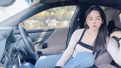 Video of a young woman with long black hair, wearing a beige top and blue jeans, seated in a car.