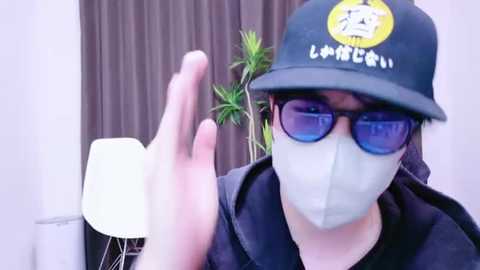 Media: Video of a person wearing a black cap with a white logo, blue sunglasses, and a face mask, waving their hand in front of a potted plant and a white chair in a room with brown curtains.